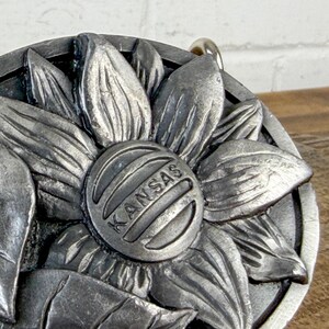 May include: A silver-coloured metal sunflower pendant. The centre of the flower features the word "KANSAS" in a circular design. The pendant has a textured, three-dimensional appearance, with visible petals and leaves.