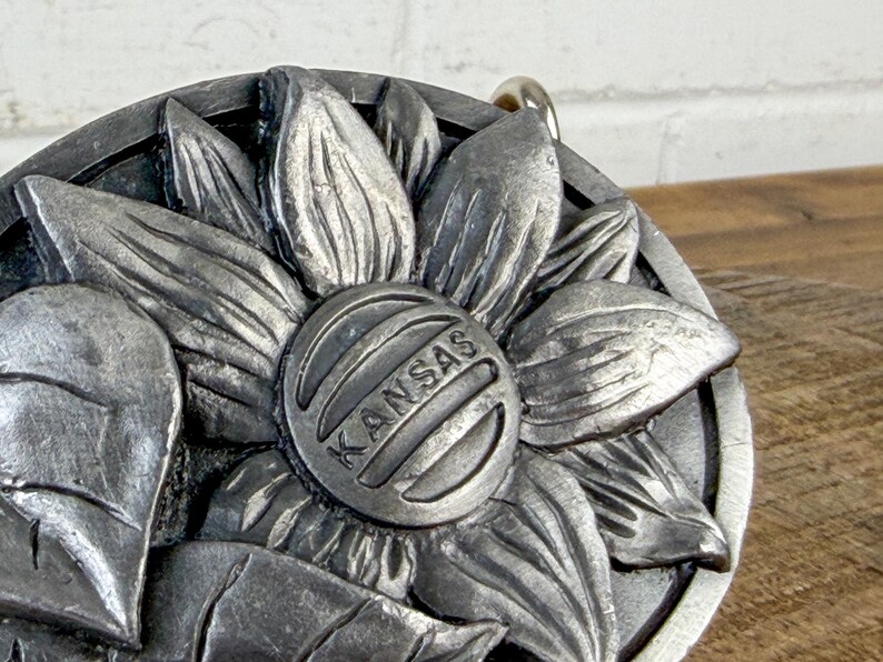 May include: A silver-coloured metal sunflower pendant. The centre of the flower features the word "KANSAS" in a circular design. The pendant has a textured, three-dimensional appearance, with visible petals and leaves.