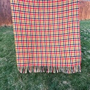 May include: A rectangular throw blanket featuring a vibrant plaid pattern in shades of red, yellow, and dark blue. The fabric displays a consistent grid design throughout the surface, and the bottom edge is finished with a row of fringe tassels.
