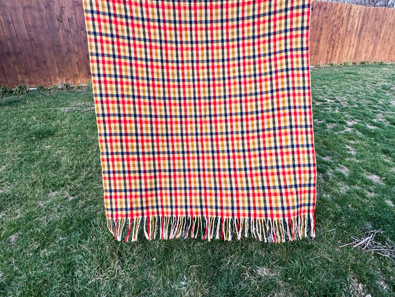 May include: A rectangular throw blanket featuring a vibrant plaid pattern in shades of red, yellow, and dark blue. The fabric displays a consistent grid design throughout the surface, and the bottom edge is finished with a row of fringe tassels.