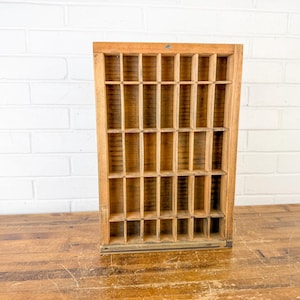 May include: A rectangular wooden printer's tray featuring a grid of numerous small, open compartments arranged in vertical and horizontal rows. The natural light brown wood grain is visible throughout the structure, which has a sturdy frame.