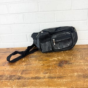 May include: A black leather fanny pack featuring multiple zippered compartments, a small flap pocket on the front, and an adjustable black nylon strap with a plastic buckle closure. The surface displays a stitched panel design throughout.