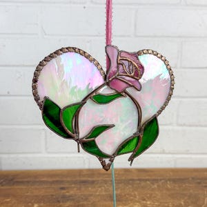 May include: A heart-shaped stained glass ornament featuring iridescent white glass panels, a pink glass rose at the top, and deep green glass leaves at the base. The pieces are joined by dark metal solder lines and it hangs from a pink ribbon.
