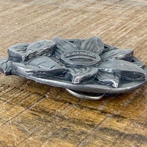 May include: A silver metal belt buckle with a floral design. The buckle features a detailed, three-dimensional flower with petals and a central ring. The buckle is set against a wooden surface.