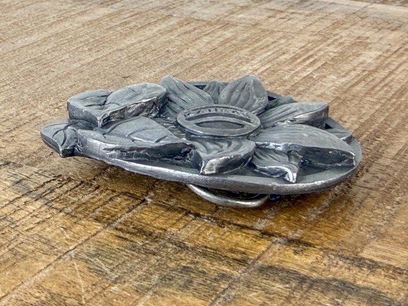 May include: A silver metal belt buckle with a floral design. The buckle features a detailed, three-dimensional flower with petals and a central ring. The buckle is set against a wooden surface.
