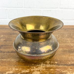 May include: A brass bowl with a wide, flared rim and a rounded, bulbous body. The metal surface displays a weathered, aged patina with visible variations in tone and texture, featuring a polished interior neck and a darker, tarnished exterior finish.