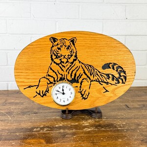 May include: Oval wooden clock featuring a black tiger illustration. The clock face is white with black numbers and hands. The wooden base has a natural wood grain finish and is supported by a dark metal stand.