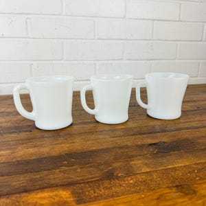 May include: Three white milk glass mugs with handles. The mugs are on a wooden surface.