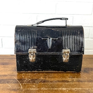 May include: A vintage black metal lunchbox with a handle and two silver latches. The lunchbox has vertical ribbed detailing on the front and top, and a decorative emblem. The lunchbox is sitting on a wooden surface.