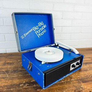 May include: A vintage Emerson Big-Big Portable Phono record player. The blue vinyl-covered case has a white turntable, a black speaker grill, and silver latches. The open lid reveals the text "Emerson Big-Big Portable Phono".