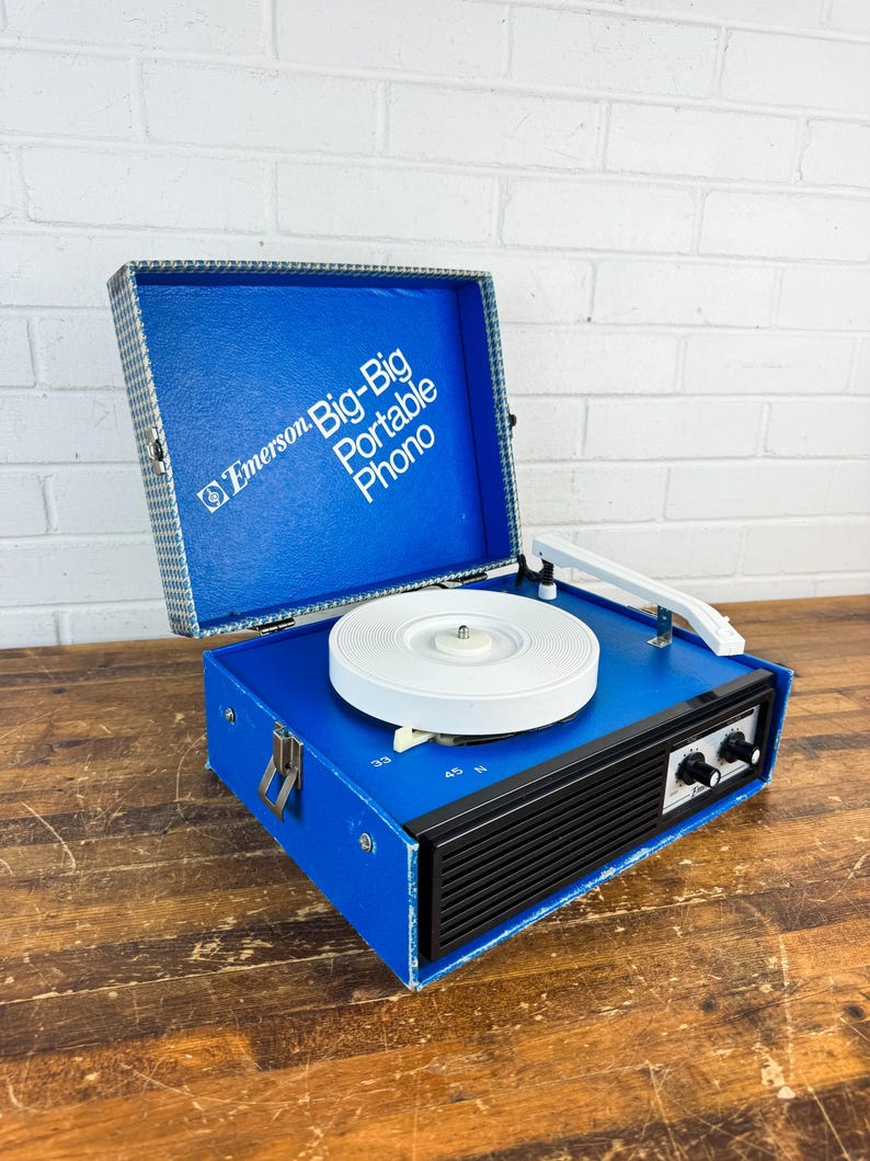 May include: A vintage Emerson Big-Big Portable Phono record player. The blue vinyl-covered case has a white turntable, a black speaker grill, and silver latches. The open lid reveals the text "Emerson Big-Big Portable Phono".