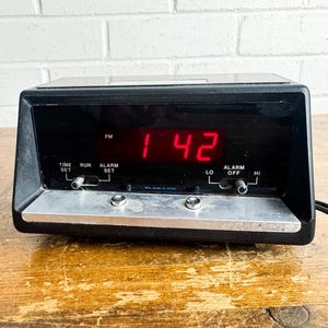 May include: Black digital alarm clock displaying the time as 1:42 PM in red numerals. The clock has a silver-colored base with buttons for time and alarm settings. The clock is on a wooden surface.