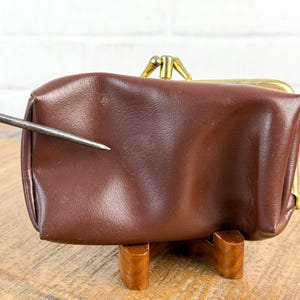 May include: A brown leather coin purse with a gold-colored clasp. The purse is rectangular and sits on a wooden stand. The leather has a slight sheen. A metal tool points towards the purse.