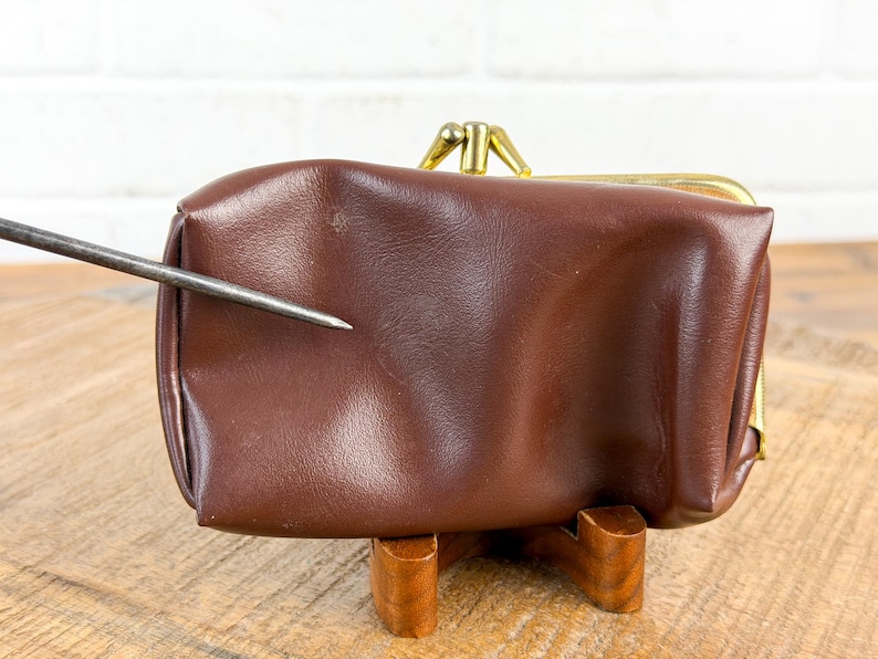 May include: A brown leather coin purse with a gold-colored clasp. The purse is rectangular and sits on a wooden stand. The leather has a slight sheen. A metal tool points towards the purse.