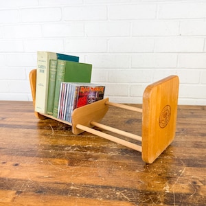 May include: A light-colored wooden book and CD holder. The holder has a rectangular shape with rounded corners and a circular design on the side. It holds several books and CDs.