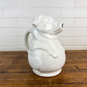 May include: A white ceramic pitcher shaped like a pig. The pitcher has a handle and a spout that resembles the pig's snout. The pig-shaped pitcher is a decorative kitchenware item.