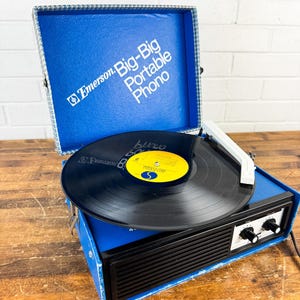 May include: A vintage Emerson Big-Big Portable Phono record player. The blue case is open, revealing a black vinyl record on the turntable. The player has a black front panel with knobs and a white tone arm. The case has the Emerson logo and the words "Big-Big Portable Phono".