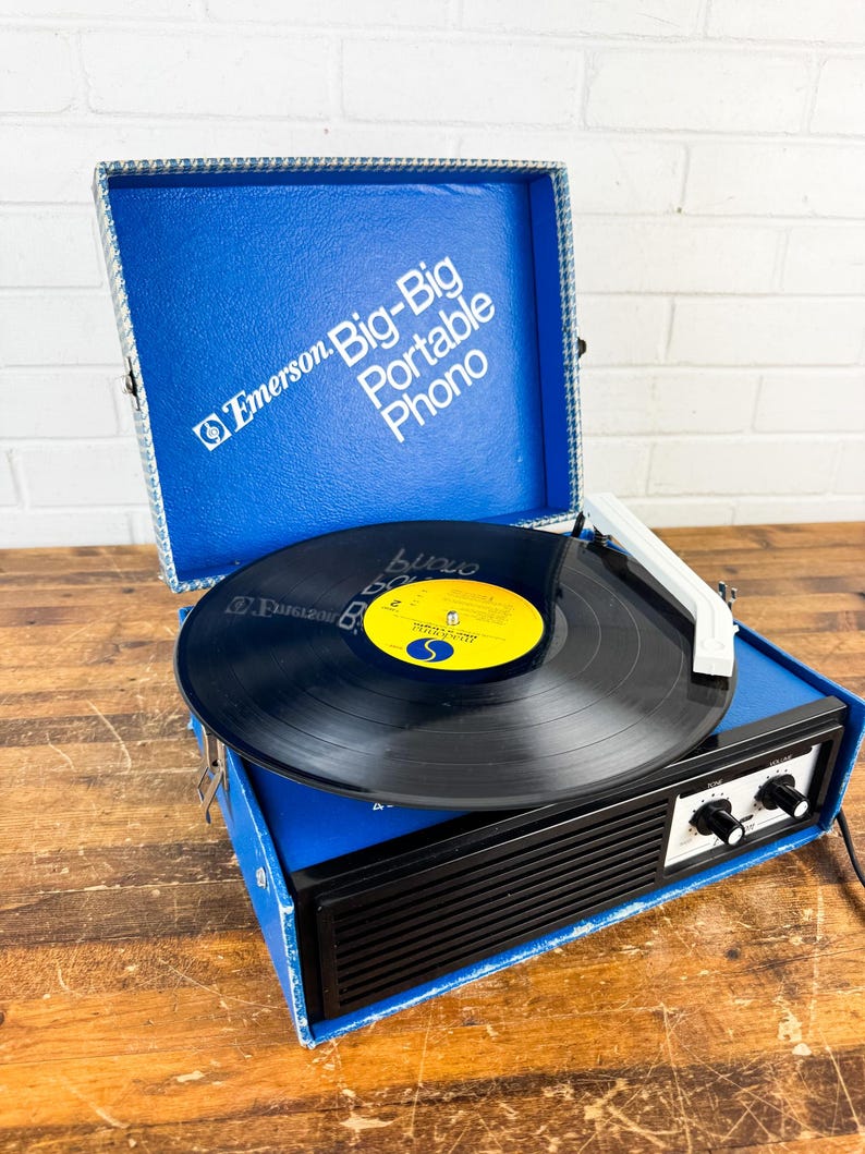 May include: A vintage Emerson Big-Big Portable Phono record player. The blue case is open, revealing a black vinyl record on the turntable. The player has a black front panel with knobs and a white tone arm. The case has the Emerson logo and the words "Big-Big Portable Phono".