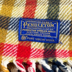 May include: A woven wool blanket featuring a plaid pattern in mustard yellow, red, white, and navy blue. A rectangular blue fabric label is stitched to the center, reading: Warranted to be a Pendleton, Pendleton Woolen Mills, Portland, Oregon.