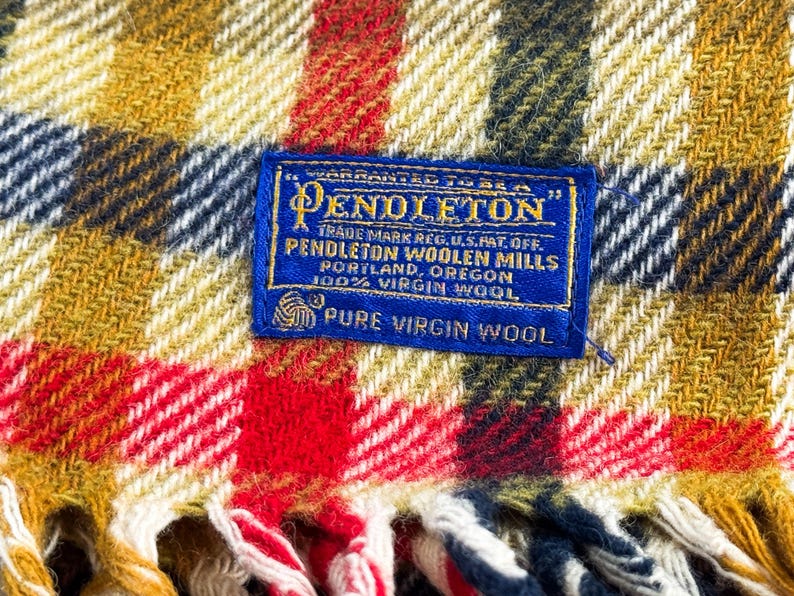 May include: A woven wool blanket featuring a plaid pattern in mustard yellow, red, white, and navy blue. A rectangular blue fabric label is stitched to the center, reading: Warranted to be a Pendleton, Pendleton Woolen Mills, Portland, Oregon.