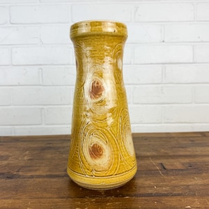 May include: A yellow ceramic vase with a flared shape and a textured surface. The vase features a pattern of swirling, light-colored lines with brown circular accents. The vase is displayed on a wooden surface.