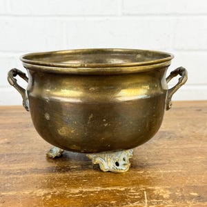 May include: A vintage brass planter with two handles and ornate feet. The planter has a rounded body and a wide rim. The brass has a warm, aged patina, giving it a classic, antique look. The planter is sitting on a wooden surface.