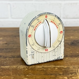 May include: A vintage off-white kitchen timer with a distressed finish and a central white dial. The face features a cream-colored circular dial with red text reading MINUTE MINDER and black numerical markings from 0 to 55 around the perimeter.