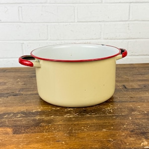 May include: A vintage cream-colored enamel pot with a red rim and handles. The pot has a white interior and sits on a wooden surface. The handles are red. The pot is a kitchenware item.