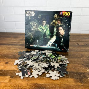 May include: A Star Wars 100-piece puzzle featuring characters from the film. The puzzle box shows Chewbacca, Princess Leia, Han Solo, C-3PO, and others. A pile of puzzle pieces sits in front of the box on a wooden surface.