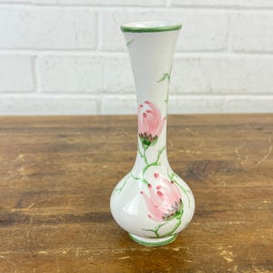 May include: A white ceramic vase with a bulbous base and a slender neck. The vase is adorned with pink floral designs and green leaves. The rim and base are trimmed with green paint. The vase is approximately 8 inches tall.