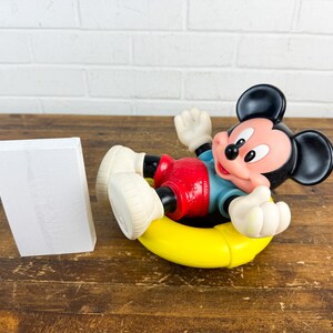 Puede incluir: Una figura de Mickey Mouse en una c&aacute;mara de aire amarilla, con pantalones cortos rojos, una camisa azul y guantes y zapatos blancos. El personaje de orejas negras est&aacute; acostado, con un objeto rectangular blanco a la izquierda.