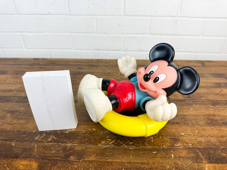 Puede incluir: Una figura de Mickey Mouse en una c&aacute;mara de aire amarilla, con pantalones cortos rojos, una camisa azul y guantes y zapatos blancos. El personaje de orejas negras est&aacute; acostado, con un objeto rectangular blanco a la izquierda.
