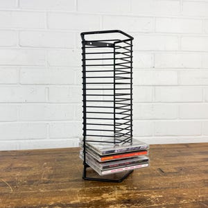 May include: A tall, black metal wire rack designed for vertical storage of compact discs. The frame features a rectangular grid structure with a flat base and a mounting bracket at the top. Three compact discs are stacked inside the bottom section.