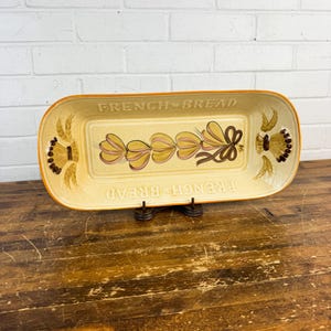 May include: Rectangular ceramic serving dish with the words "FRENCH BREAD" in orange lettering. The dish has a cream-colored background with a floral design in yellow and brown. The dish is on a dark metal stand.