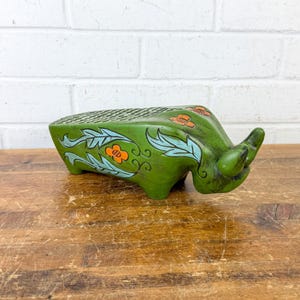 May include: Green ceramic bull-shaped planter with orange and blue floral accents. The planter has a textured top and is set on a wooden surface. A decorative piece for home or garden.