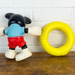 Puede incluir: Una figura de juguete vintage de Mickey Mouse con cabeza y orejas negras, camisa azul, pantalones cortos rojos y guantes y zapatos blancos. Un juguete de anillo amarillo est&aacute; al lado de la figura. Los juguetes est&aacute;n sobre una superficie de madera.