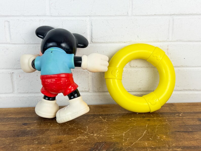 Puede incluir: Una figura de juguete vintage de Mickey Mouse con cabeza y orejas negras, camisa azul, pantalones cortos rojos y guantes y zapatos blancos. Un juguete de anillo amarillo est&aacute; al lado de la figura. Los juguetes est&aacute;n sobre una superficie de madera.