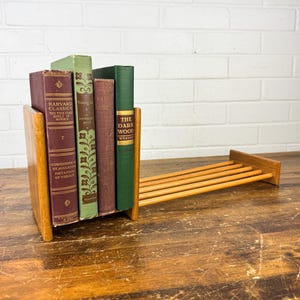 May include: A wooden bookend with a built-in shelf holds several vintage books. The books have maroon, green, and brown covers, with titles like "Harvard Classics" and "The Dark Wood." The bookend and shelf are made of light-colored wood.