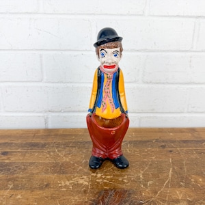 May include: A vintage ceramic clown figurine. The clown wears a black bowler hat, a yellow jacket with blue trim, and red pants. The clown has a painted face with blue eyes and a red mouth. The figurine is standing on a wooden surface.