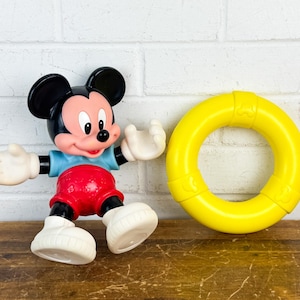 Puede incluir: Una figura de Mickey Mouse con un salvavidas amarillo. La figura de Mickey Mouse lleva una camisa azul, pantalones cortos rojos y zapatos blancos. El salvavidas es de color amarillo brillante.