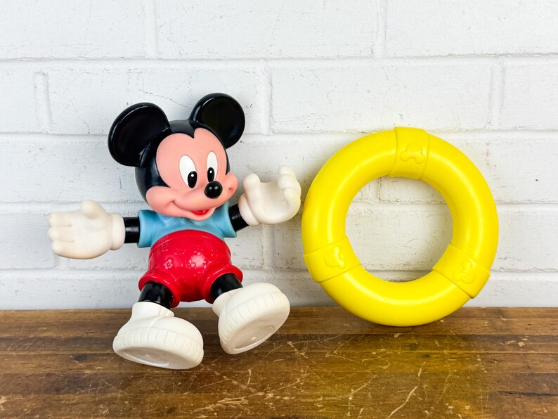 Puede incluir: Una figura de Mickey Mouse con un salvavidas amarillo. La figura de Mickey Mouse lleva una camisa azul, pantalones cortos rojos y zapatos blancos. El salvavidas es de color amarillo brillante.