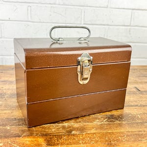 May include: A brown metal toolbox with a silver handle and clasp. The rectangular box has a shiny finish and is sitting on a wooden surface. The toolbox is designed for storing tools and hardware.