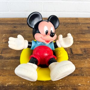 Puede incluir: Una figura vintage de Mickey Mouse. La figura tiene orejas negras, una cara rosada y guantes y zapatos blancos. Mickey lleva una camisa azul, pantalones cortos rojos y est&aacute; sentado sobre una base amarilla. El juguete es de pl&aacute;stico.