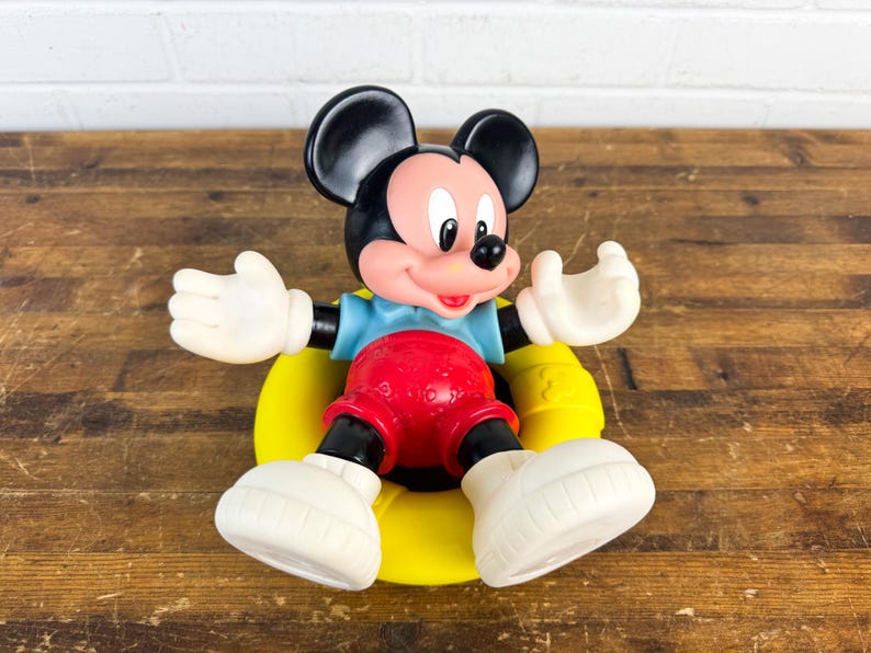 Puede incluir: Una figura vintage de Mickey Mouse. La figura tiene orejas negras, una cara rosada y guantes y zapatos blancos. Mickey lleva una camisa azul, pantalones cortos rojos y est&aacute; sentado sobre una base amarilla. El juguete es de pl&aacute;stico.