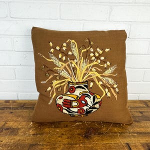 May include: Brown square pillow with an embroidered design. The design features a vase with a bird and floral pattern, holding wheat-like stalks. The pillow is placed on a wooden surface against a white brick wall.