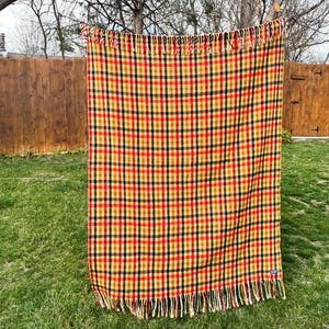 May include: A rectangular woven blanket featuring a vibrant plaid pattern in shades of mustard yellow, deep red, and dark navy blue. The textile has a consistent grid design throughout and is finished with long, straight fringe along the top and bottom edges.