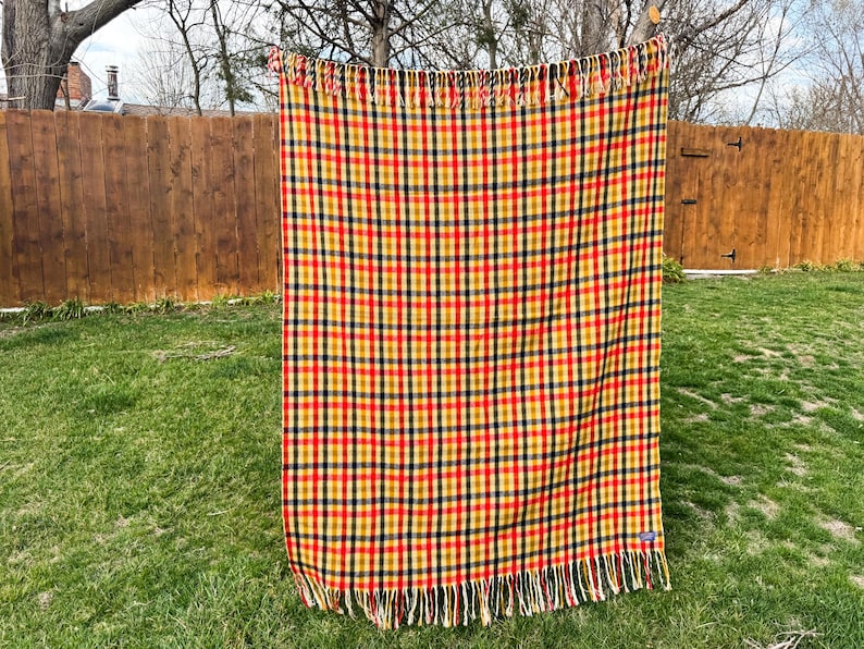 May include: A rectangular woven blanket featuring a vibrant plaid pattern in shades of mustard yellow, deep red, and dark navy blue. The textile has a consistent grid design throughout and is finished with long, straight fringe along the top and bottom edges.