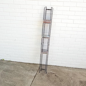 May include: A tall, narrow, black metal wire rack designed for storing compact discs. The vertical storage unit features numerous horizontal slots for organizing individual cases, with a few discs currently placed in the upper and middle sections.