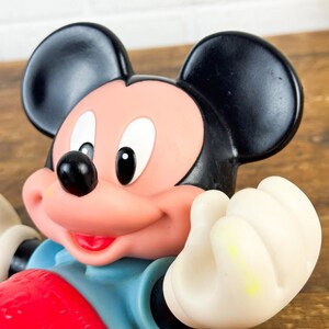 Puede incluir: Primer plano de una figura de Mickey Mouse. La figura tiene orejas negras, cara rosa y nariz negra. Lleva una camisa azul y pantalones cortos rojos. Las manos son blancas y la figura sonr&iacute;e.