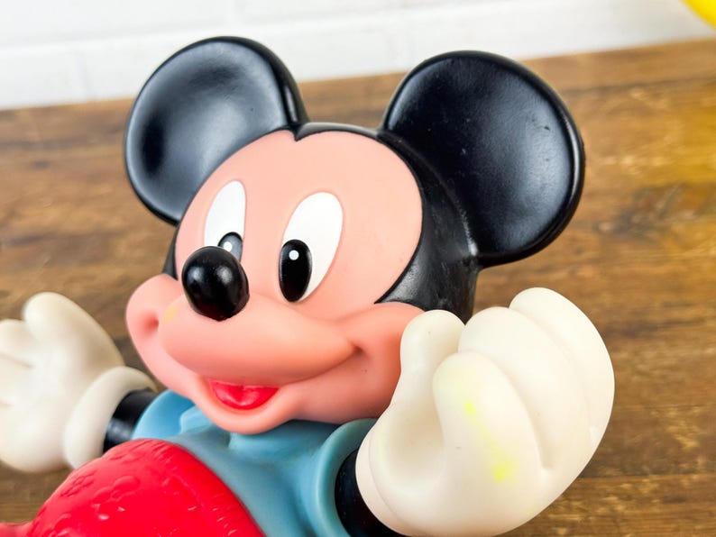 Puede incluir: Primer plano de una figura de Mickey Mouse. La figura tiene orejas negras, cara rosa y nariz negra. Lleva una camisa azul y pantalones cortos rojos. Las manos son blancas y la figura sonr&iacute;e.